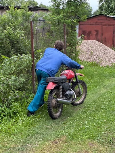 A person in a blue jacket and teal pants is pushing a red dirt bike on a grassy area. The person is facing away from the camera, with their right hand on the handlebars. The dirt bike has a black seat and silver exhaust pipe. In the background, there is a chain-link fence, a wooden fence, a pile of gravel, and a red garage. The scene appears to be in a rural or semi-rural setting, possibly in Piriutino, Russia, given the presence of a typical Russian garage and the overall atmosphere. The lighting suggests an overcast day.