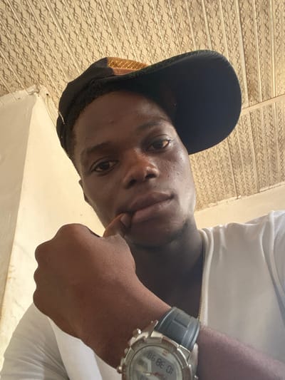 A close-up, low-angle photograph captures a single male individual indoors. The subject has dark skin and is wearing a white short-sleeved t-shirt and a black baseball cap with a patterned, possibly gold and green, brim, worn backward. A thin gold-colored chain is visible around his neck. His left hand is raised, with the index finger touching his lower lip/chin area, while the thumb rests beneath his chin. A silver-toned wristwatch with a dark grey rubber strap is present on his left wrist, displaying a digital reading partially visible as "10 38 98". The background features a light-colored, possibly cream or beige, wall to the left and a patterned ceiling composed of repetitive, interlaced panels, extending across the top and right side of the frame. The subject is looking directly into the camera. The precise location within the city of Igbesa, Nigeria, cannot be determined from the visual evidence presented.