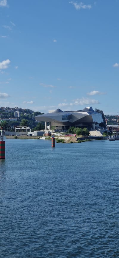 The image displays an exterior, daylight scene featuring a prominent modern architectural building situated on the bank of a wide river, observed from the water. The location is in the city of La Mulatière, France.
The central subject is the Musée des Confluences, characterized by its complex, angular, and multi-faceted design. The building's exterior is composed of reflective metallic paneling and large glass sections, creating an irregular and dynamic form. It is positioned on the river's edge, with its elevated sections supported by concrete structures.
The lower half of the photograph is dominated by the river, exhibiting deep blue-green water with numerous small ripples across its surface. Two navigational buoys are visible in the river. The buoy on the left features horizontal green and red stripes, topped with a white rectangular sign displaying the text "VHF" above the number "18". The buoy on the right also has horizontal green and red stripes, topped with a smaller white sign showing the number "15".
The riverbank directly in front of and to the right of the museum features a narrow strip of land with sparse green vegetation, including several small trees. A dirt path is discernible along this bank, on which several indistinct human figures are observed walking or standing. A large pink banner or installation is visible on the bank, displaying partially obscured text that reads "ONLY LYON".
In the background, behind the museum and to its left, a verdant hillside rises, densely populated with numerous residential buildings of various sizes and architectural styles, interspersed with extensive tree coverage. A road with a few vehicles is visible at the base of the hillside, near the museum's foundation.
The upper portion of the image shows a clear, bright blue sky with scattered, small white cumulus clouds, which are slightly more concentrated above the distant hillside. The overall lighting suggests a sunny day.