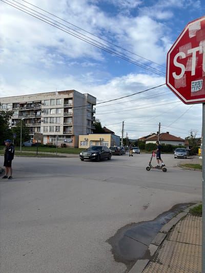 The image displays an active street scene in Slivnitsa, Bulgaria, under a bright, partly cloudy daytime sky. The perspective captures elements from the sky to the road, featuring buildings and individuals.
In the upper left, a prominent red octagonal stop sign is visible, bearing the white Cyrillic word "СТОП" (STOP). A white sticker with "САНИ ГЕРО" is affixed to the sign. Overhead, multiple power lines crisscross the blue and white sky, connected to utility poles.
On the asphalt road, a young person, dressed in a dark t-shirt and shorts, is actively riding an electric scooter, positioned in the mid-ground. Their body is angled, demonstrating balance. Another person, wearing a dark jacket, shorts, and a blue cap, stands near the bottom edge of the frame, partially visible on the sidewalk or road edge.
Several vehicles are present, including a dark sedan parked on the right side of the road further back, and a silver car visible in the distance. Along the right side, a paved sidewalk features distinctive yellow tactile paving for the visually impaired. A large, dark puddle of water lies conspicuously on the roadside next to the sidewalk, suggesting recent rain or standing water.
To the left, a multi-story apartment building with numerous windows and balconies stands, showing signs of wear. Adjacent to it, a smaller, yellow commercial building with an air conditioning unit and some indistinct signs is visible. Further down the street on the right, smaller residential houses with tiled roofs can be seen. The scene depicts a typical urban or suburban environment with a mix of residential and commercial structures.