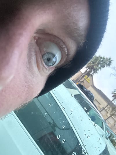 A forensic close-up image captures the left eye of an individual with a blue-grey iris, looking forward. The subject's skin is fair, and a black beanie or similar dark head covering is visible above the eye. The foreground features the exterior of a vehicle's side window, which is covered with numerous clear water droplets, indicating recent precipitation. Through the rain-speckled window, a light-colored building with a distinctive peaked roofline is visible in the background. A prominent yellow sign on the building displays partial text, including "OLD" and "OYAL," consistent with "The Old Royal Palace" building. Multiple palm trees are present against an overcast sky. The scene is located in the vicinity of 815 N La Brea Avenue, Inglewood, California, United States.