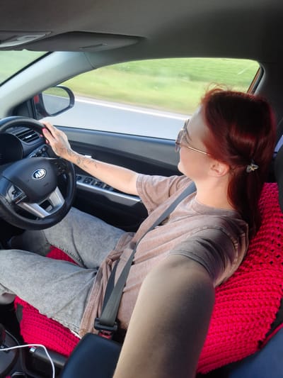 A person with red hair, wearing sunglasses and a beige t-shirt, is driving a car. They have tattoos on their left arm and hand, which is on the steering wheel. The car's interior is visible, including the dashboard and a bright pink knitted seat cover. The background shows a blurry green landscape, suggesting the car is in motion on a road. The image does not provide any information about the specific location in the city of Поварёнки, Russia.