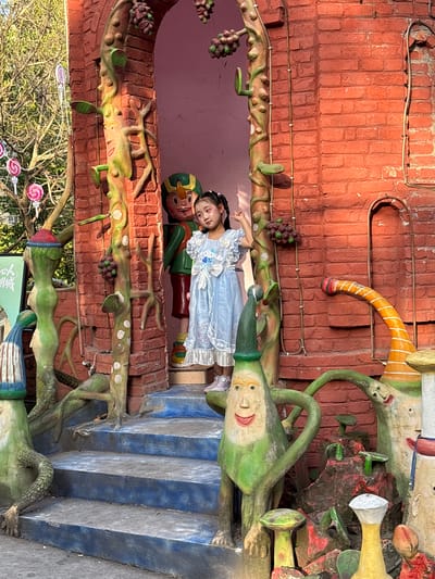 The photo shows a young girl in a light blue dress lying down next to a Pinocchio statue. They are positioned within an elaborately decorated archway or entranceway. The structure features brickwork, sculptural vines with grapes, and green, stylized, cartoonish figures.

The location is Baqiao, China.