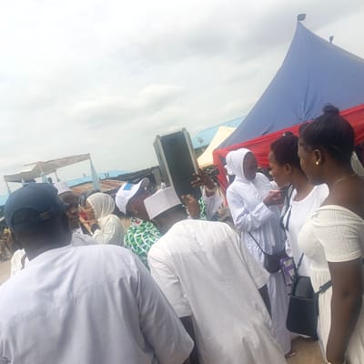 Here's a detailed description of the image:
**Setting:** The image appears to be taken outdoors, likely in Gudi, Nigeria, based on the request. The sky is overcast. There are tents visible in the background, one with blue and red fabric. The ground is paved.
**People:** There is a group of people gathered, predominantly dressed in white clothing, suggesting a special occasion. The group contains both men and women. Some of the women wear hijabs. One woman has a bare shoulder dress. A man in the foreground wears a dark baseball cap.
**Objects:** A large speaker is positioned prominently. The women are carrying various small purses.
**Actions/Interactions:** The people are engaged in conversation and seem to be socializing. Some are gesturing with their hands.
**Overall Impression:** The scene suggests a gathering, possibly a celebration or ceremony, taking place outdoors in Gudi, Nigeria. The atmosphere appears relaxed and social.