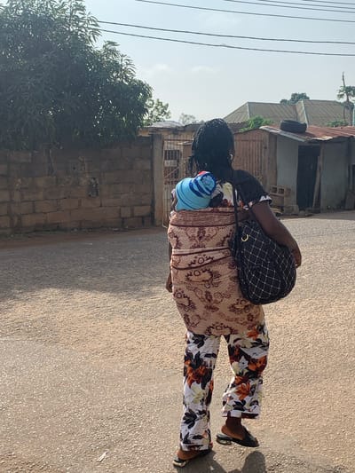 The image depicts a person walking on a paved road in what appears to be a residential area in Jos, Nigeria. The person is carrying a baby on their back, covered with a blue and white striped hat and a patterned fabric. They are wearing a long-sleeved, patterned top and floral-patterned pants. A black and white checkered bag is slung over their shoulder.
The scene is outdoors, with buildings and trees visible in the background. The buildings have corrugated metal roofs and brick walls, suggesting a warm climate. Power lines are also visible, running across the sky. The time of day appears to be late afternoon, as indicated by the shadows cast on the ground. The weather seems to be clear and sunny.
There is no visible text in the image. The overall impression is of an everyday scene of a person going about their daily activities, likely in a bustling urban or semi-urban environment. The focus is on the person and the baby, highlighting the act of carrying and caring for a child in a public space.