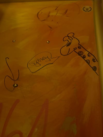 The image shows a close-up of a wall with yellow and orange paint strokes. There are several distinct drawings and markings on the wall. On the upper right, there is a circular emblem with laurel leaves surrounding a stylized cross. To the left of the emblem, faint dark lines suggest a larger, unfinished drawing, possibly of a face or figure. In the center of the image, a cartoon-like drawing of a giraffe's head and neck is rendered in black outline. The giraffe has large eyes and a spotted pattern on its neck. To the left of the giraffe, a speech bubble with the name "Sydney" written inside it is drawn in black outline. Below and to the left of the speech bubble, a thin, curved black line forms a shape resembling a loop or a partial question mark. Several small, circular metal objects, possibly rivets or studs, are embedded in the wall, reflecting light. Faint red graffiti is visible towards the bottom of the image. The overall impression is of a child's drawing or markings on a painted surface. There is no discernible information within the image to suggest a specific location in Leipzig, Germany, or any details about people, actions, or interactions.