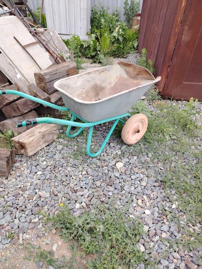 A weathered, light gray wheelbarrow with a worn, light orange wheel rests on a bed of gravel and sparse green vegetation. The wheelbarrow's turquoise handles are visible, with black tape wrapped around the ends. Behind the wheelbarrow, a stack of rough, weathered wooden planks and logs lies on the left. In the background, a light-colored wooden fence or structure is partially visible, along with dense green foliage and a large, reddish-brown corrugated metal sheet leaning against something unseen. The scene appears to be an outdoor storage area, possibly in a rural or industrial part of Ekibastuz, Kazakhstan, given the presence of construction materials and the general state of disrepair. There are no people present.