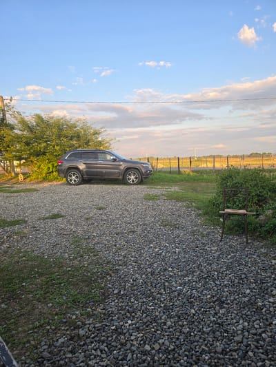 A dark gray Jeep Grand Cherokee is parked on a gravel driveway next to a dense green tree. The driveway is covered in small, dark gray and tan pebbles. To the left of the car, the tree's foliage is thick and lush, with sunlight filtering through its branches. In the distance, a fence line runs horizontally across the frame, with a field of vegetation beyond it. The sky is a bright blue with scattered white and wispy clouds, tinged with warm hues of orange and pink, suggesting it's either early morning or late afternoon. A thin black wire stretches across the sky, likely a utility line. A metal chair with wooden slats is placed near some dense green bushes in the background. The overall setting appears to be a rural or semi-rural area in Ingiri, Georgia. There is no visible text in the image.