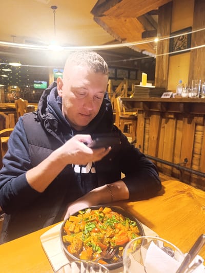 A person sits at a wooden table inside a restaurant. The person is a light-skinned man with short, light-colored hair. He is wearing a dark hoodie with a black vest over it. He holds a mobile phone in his hands, appearing to be focused on it. In front of him is a dark skillet filled with what appears to be a cooked vegetable dish, garnished with green herbs. Next to the skillet is a glass containing napkins and cutlery. The restaurant has a rustic, wooden interior with a wooden bar and other wooden furniture visible in the background. A blurry view through a window shows some exterior lights. The location is Chernogolovka, Russia.