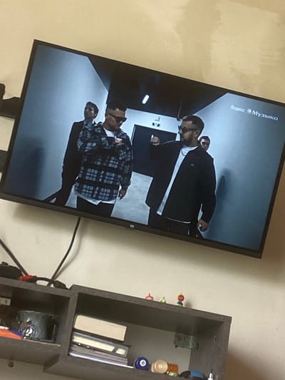 The image displays a television screen showing a music video with two men in the foreground and a third person in the background. The man on the left is wearing a plaid shirt over a white t-shirt and dark pants, with sunglasses. The man on the right is wearing a black jacket over a white t-shirt and dark pants, also with sunglasses. The television is mounted on a wall above a shelf unit. On the shelf, there are books, a mug, and various small objects. The overall scene suggests an indoor domestic setting, likely a living room. The location of Kisliakovskaya, Russia, is mentioned in the prompt but is not visually discernible from the image itself.