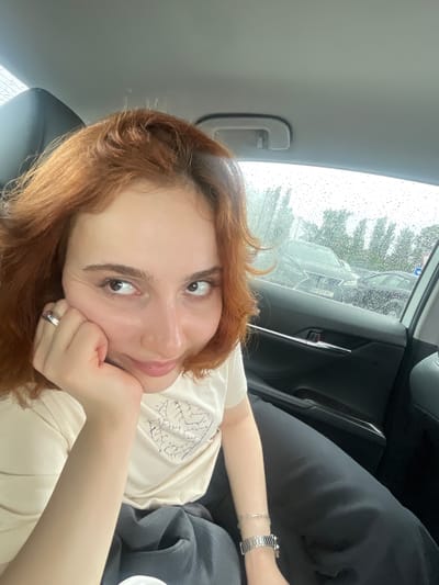 A young adult female with red hair is lying on a dark car seat inside a vehicle. She's resting her head on her hand and looking at the camera. She's wearing a light-colored t-shirt and a silver wristwatch and bracelet on her left wrist. The car window shows rain droplets and a blurry view of other cars and trees outside. Based on the license plate visible on the car outside, the location is likely in Georgia.