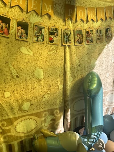 The image depicts a festive scene, likely a birthday celebration for a child, with a backdrop of a yellow banner displaying the words "ПРИ-РОЖДЕНИЕ" (birth) and a string of photos from birth up to 10 months. The photos are small, framed tags, each with a numerical label in Russian indicating the month. The backdrop itself has a sandy texture with imprints of seashells.
In the foreground, to the right, is a cluster of balloons, predominantly light blue and teal, with one large, elongated teal balloon forming the shape of the number '1'. There are also some brown and white balloons. Partially visible in the lower left is a yellow and green inflatable toy, possibly a sea creature like a turtle. The lighting suggests an indoor setting with natural light casting shadows from the right.
This scene is likely located in Lyamino, Russia, given the presence of Russian text on the banner and photo tags, and it appears to be a decorated room prepared for a child's birthday party. There are no identifiable people actively engaged in the frame; the focus is on the decorations and celebratory items.