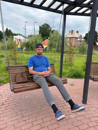 A man is sitting on a wooden swing in an outdoor park area. He is wearing a blue t-shirt, grey sweatpants, black socks with white stripes, and blue Crocs with red and white stripes. He has a dark baseball cap on his head, worn backward. His hands are clasped in his lap, and his eyes are closed. A brown purse is on the seat to his left. The swing is suspended by metal chains from a dark metal frame. The ground is paved with red bricks. In the background, there is a grassy area with scattered white flowers and trees. Behind the trees, a red-brick house with a gabled roof and chimneys is visible. To the left of the house, a playground structure resembling a ship's mast can be seen, along with red flags. The sky is overcast. The location is the park in Breytovo, Russia.