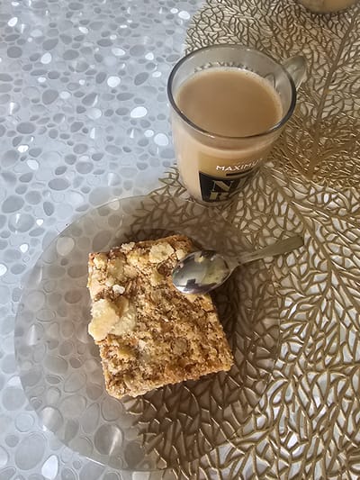 The image shows a close-up, top-down view of a square piece of pastry on a clear, patterned plate, next to a glass mug filled with a light brown beverage. A silver spoon rests on the plate, partially on the pastry. The mug has a label that reads "MAXIMUS". The setting appears to be a table covered with a textured white placemat and a gold, leaf-patterned placemat beneath the mug. There is no indication of the city of Таёжное, Russia, or any specific location within it. The scene is static, with no people or discernible actions taking place.
