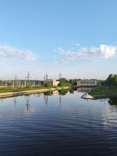 The image captures a waterway in 3-Y Uchastok, Russia. On the left bank, a large electrical substation is visible, with numerous power lines and transformers. These structures are reflected in the water. Further along the bank, a building is nestled among greenery. The opposite bank features a building with a distinctive architectural design. A small, white structure juts into the water, separated from the bank by a fence. The sky is mostly clear with scattered clouds. The water surface is rippled, reflecting the sky and the shoreline.
