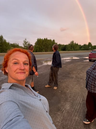 In this outdoor selfie, a woman with vibrant red hair and striking blue eyes smiles at the camera. She is wearing a light blue button-down shirt. Behind her, a group of people are visible. One man in a dark jacket and brown pants walks away from the camera, while another man in a plaid shirt stands further in the background. The scene appears to be a parking lot after a rain, with puddles reflecting the overcast sky. A faint rainbow is visible in the upper left corner of the image. The presence of trees and the overall atmosphere suggest a rural or suburban setting, consistent with the provided location context of Sovkhoznyi, Russia. The soft lighting and the rainbow hint that the time of day is likely late afternoon or early evening, possibly following a shower. The mood is casual and relaxed, with the subjects appearing to be enjoying an outdoor moment together. No visible text is present in the image.
