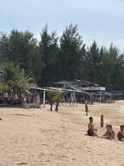 The image depicts a beachside scene in Meunasahbale, Indonesia, characterized by a sandy shore backed by dense pine trees. In the foreground, several young boys, some with sandy skin, are seated on the beach. Further back, a variety of structures line the edge of the sand, including open-air seating areas with tables and chairs, some decorated with string lights. A multi-story building with balconies is visible in the background, suggesting a restaurant or resort. People are seen sitting at tables, walking, and interacting in these shaded areas. The overall atmosphere appears to be a casual, recreational setting on a cloudy day.