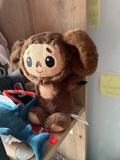 A close-up shot shows a brown plush monkey toy lying on its side in a wooden box. The monkey has a round face with large, expressive eyes, a small nose, and a smiling mouth. Its fur is soft and fluffy, and its limbs are relaxed. Other objects are visible in the box, including a small blue shark plush toy with its tail visible, a red clip, and a metal coin. There are also some colorful pipe cleaners and a white tag with text that appears to say "CE MADE." A piece of yellow paper with handwritten text is attached to the wall behind the box, and another yellow note is visible to the left. The lighting suggests an indoor setting, possibly a room or storage area. The overall scene appears to be a collection of toys and small items in a disorganized yet homey environment.