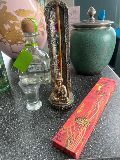 The image presents a collection of objects arranged on a dark, speckled surface. These objects include:
* A turquoise ceramic jar with a silver lid and a blue knob on top.
* A red box of "The Mother's India Fragrances" incense sticks, specifically "Rishi" scent.
* An incense burner with a small Buddha figure and a row of colorful gemstones. An incense stick sits in the holder.
* A Patron tequila bottle.
The overall impression is that of a collection of spiritually-themed and decorative items, casually arranged on a surface. The image doesn't suggest a specific location in Liverpool, UK. There are no people, actions, or interactions evident in the image. The scene is a still life of assorted objects.