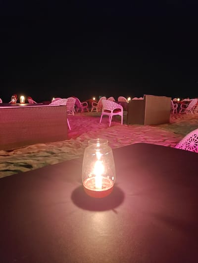 The image depicts an outdoor dining area, likely on a beach, at night. The location is potentially in Arambol, India. The scene is illuminated primarily by candlelight, giving the image a warm, pinkish hue. In the foreground, there is a candle inside a glass jar on a dark, possibly leather-like, table. The candle is lit, and its light reflects on the table surface. In the background, there are several tables and chairs, mostly made of rattan or similar wicker material, scattered across what appears to be sand. Other candle lanterns are visible on the tables in the background. The environment appears to be a beachside setting, with no clear indication of buildings or urban structures visible. The sky is dark, suggesting it is night, with a single star visible. There is no apparent human activity in the image.
