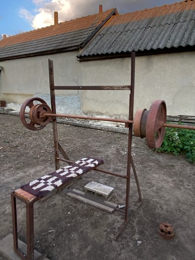 The image shows an outdoor workout bench with a barbell and weights, situated in a yard with a dirt ground. The bench is made of rusted metal and has a small, patterned mat on the seat. The barbell is loaded with two large, circular weights on each side. In the background, there is a building with a tiled roof and a stucco wall. To the right of the bench, there is green foliage. The sky above is partly cloudy, with the sun casting a warm light on the scene. There are no people visible in the image. The scene appears to be in a rural or semi-rural setting. This image was taken in Cairaclia, Moldova.