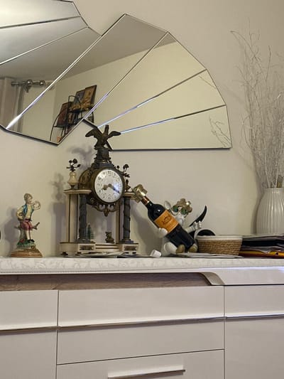 This image depicts a static indoor scene in Colombes, France, focusing on a decorated white dresser or credenza. Above the dresser, a large, segmented mirror with a fan-like design is mounted on a light-colored wall. The mirror reflects a portion of the room, including light-colored curtains on the left and a shelf unit displaying several framed pictures or icons on the opposite wall.
On the dresser's top surface, which is covered by a white, subtly patterned runner, various decorative objects are arranged. From left to right:
1. A ceramic figurine of a boy in a blue shirt and red shorts, seated on a green and brown tree stump, playing a flute.
2. An ornate, antique-style mantel clock with a dark bronze or brass finish. It features a bird (likely an eagle or goose) atop its structure, which includes a white clock face with dark Roman numerals, flanked by two light-colored pillars. Below the clock face, miniature decorative elements, including a small green Christmas tree and another tiny figurine, are visible.
3. A white figurine, resembling a cartoon character with sunglasses, acts as a wine bottle holder. It holds a dark glass wine bottle with an orange label featuring "CHATEAU DE LA RONCIERE" text and an illustration. The bottle's neck is adorned with a gold-colored stopper.
4. A small, round woven basket with a light-colored top rim.
5. A stack of papers or documents located to the right of the basket.
6. A white, fluted ceramic vase containing several slender, silver-colored, glittery decorative branches.
The dresser itself is white with horizontal accent lines, indicating multiple drawers or cabinet doors. No individuals are present in the frame.