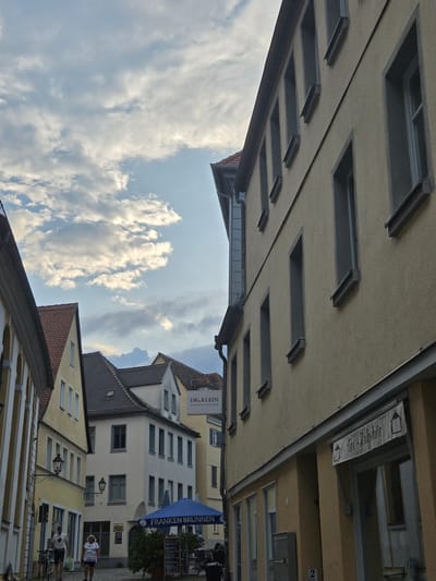 This is a photograph of a street scene in Ansbach, Germany, taken from a low angle looking upwards. The buildings are old and have traditional architecture, with tiled roofs and cream-colored facades. The sky is filled with fluffy white clouds.
In the foreground, there are several modern awnings extending from the buildings. One of these awnings is blue and has text that reads "FRANKEN BRUNNEN". Another awning has the text "DR. KLEIN" and "Die Partner für thee Finanzen". There is also a banner that appears to say "Tec-Zubhör" with a graphic of a tea cup.
In the background, there are people walking on the street, suggesting it is daytime and likely a busy period. One person is wearing a white t-shirt with the word "HELLO" and "KISS" on it. There are bicycles parked outside one of the buildings. The lighting suggests it is a bright, possibly sunny day, but the presence of many clouds indicates overcast conditions or a partly cloudy sky. The overall impression is a charming European town with a mix of old and new elements.