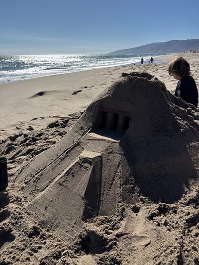 On a sunny day at a beach in Malibu, California, a child, viewed from behind, is focused on a large, intricately constructed sandcastle. The sandcastle, with its tiered base and a top section resembling a fortress with three arched openings, dominates the foreground. The beach stretches out towards the ocean, where gentle waves are breaking on the shore. In the distance, the Pacific Ocean reflects the bright sun, and a hazy mountain range forms the backdrop. Other beachgoers are visible in the distance, some walking along the water's edge, adding to the serene beach scene.