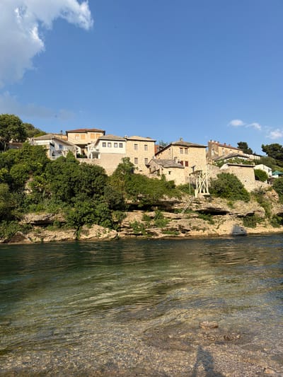 The image shows a picturesque scene in Mostar, Bosnia and Herzegovina. A row of traditional stone buildings with terracotta roofs is perched on a steep, rocky cliffside overlooking a clear, shallow river. Lush green trees and vegetation cling to the cliff, interspersed with the historic architecture. One of the buildings features a distinctive wooden structure, possibly a viewing platform or an old water mill mechanism, with stairs leading up to it. The river, with its pebbles visible through the water, reflects the sunlight and the surrounding greenery, creating a serene and inviting atmosphere. The sky is a vibrant blue with a few scattered white clouds, suggesting a clear and pleasant day, likely in the late morning or early afternoon due to the bright sunlight casting shadows. The overall mood is peaceful and tranquil, showcasing the timeless beauty of the historic town. There is no visible text in the image.