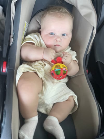 A high-angle shot captures a baby lying in a car seat. The baby is wearing a light yellow outfit and white socks. The baby's hands are holding a colorful rattle toy. The car seat is tan and black. The scene is likely inside a vehicle.