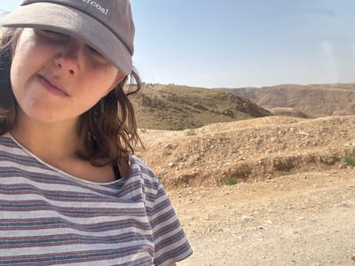 A female subject is partially depicted in a close-up selfie-style photograph taken in an arid outdoor environment. She wears a grey baseball cap with "charcoal" embroidered in white on the front, casting a shadow over her upper face. Her brown, wavy hair is visible below the cap. She has a small, silver-colored leaf-shaped earring on her right earlobe. Her attire includes a light-colored t-shirt with thin horizontal stripes in red and blue; a black garment strap is visible beneath her shirt on her right shoulder. She appears to be looking towards the camera, with a slight upward tilt to her head. The background consists of a vast, sunny desert landscape characterized by numerous rolling, parched hills and mountains under a clear, light blue sky. The immediate ground is covered in light-colored gravel and sparse dry vegetation. This scene is located in the Megilot Yam HaMelakh Regional Council, Palestinian Territory.