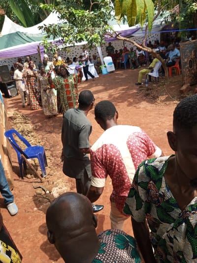 This image depicts an outdoor social gathering or celebration in Oko Town, Nigeria, under bright daylight conditions. The ground is reddish-brown earth, with scattered small rocks and a distinct pile of soil or debris along the left foreground. Overhead, green tree foliage provides partial shade, and a light-colored canopy with green and purple fabric drapings is set up in the background, alongside another similar canopy.
In the immediate foreground, four men are partially visible from behind. From left to right: a man with a shaved head wearing a green and white patterned shirt; a man with short dark hair in a plain dark grey tunic; a man with short dark hair in a red and white patterned shirt with a central plain white panel; and on the far right, the side of a man's head and part of a green, white, and red patterned shirt.
In the midground, a line of at least seven women in colorful, patterned traditional Nigerian dresses (ankara style) are moving towards the left. The prominent woman in the foreground of this group wears a green and orange patterned dress, has her arms slightly raised, and appears to be holding purple fabric. To the left of this group, a blue plastic chair lies on its side, near a wooden or metal stick. On the right midground, a large, dark, possibly wooden speaker box stands.
In the background, numerous individuals are gathered under the canopies. Many are seated on plastic chairs (white, green, red, blue), while others are standing. Tables draped with white tablecloths are visible. One man in a light green traditional outfit is seated on a white chair on the right side of the gathering, near a small bottle on the ground. Another man wearing a light blue shirt is seated further to the right. The overall scene suggests a festive or ceremonial event.