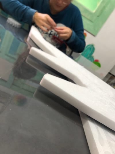 A blurry image depicts a person wearing a blue long-sleeved shirt, using their hands near an indistinct object. In the foreground are white, foam-like letterforms on a dark gray surface. The background shows a partially visible wall with a green frame, a wall painting with a cactus, and other out-of-focus elements. No specific information indicates the location is in Beccar, Argentina.