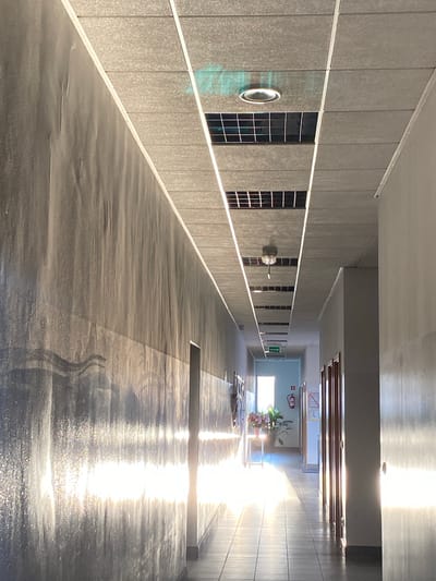 The image shows a well-lit hallway. The ceiling is made of acoustic tiles and features recessed lighting and ventilation grates. A large mural or decorative covering runs along the left wall, reflecting the sunlight streaming in from the right side of the hallway. The right side of the hallway is lined with doors. At the end of the hallway, a window allows natural light to enter, illuminating a floral arrangement on a stand and a fire extinguisher mounted on the wall. A green directional sign with an arrow is visible on the ceiling. The floor is tiled. The overall impression is one of a clean, modern, and functional space, possibly an office building or institutional setting. This specific location is not identifiable from the image alone without additional information.
