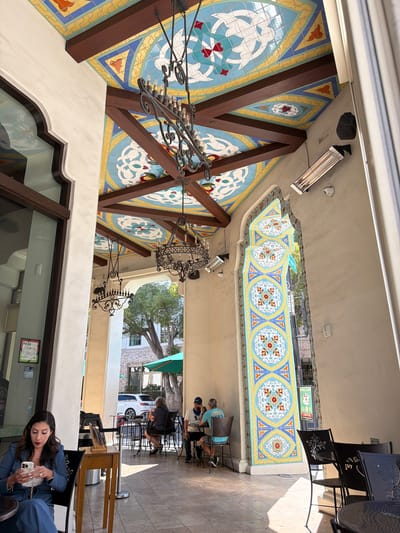 The image depicts an outdoor seating area of a cafe, likely located in Pasadena, United States of America, characterized by its Mediterranean-inspired architecture. The ceiling is adorned with vibrant, colorful tiles in geometric patterns and motifs of birds and hearts, supported by dark wooden beams. Two ornate, wrought-iron chandeliers hang from the ceiling. The walls are a neutral tone, with arched openings and decorative tiled panels.

In the foreground, a woman in a blue pantsuit is seated at a table, looking at her phone. To her right, a wooden table displays a few items. Further into the scene, several other patrons are seated at tables. Two men are engaged in conversation at one table, and a woman is seated at another. Beyond the seating area, a white SUV is parked on the street, and trees and buildings are visible in the background. The ground is paved with stone tiles, and sunlight casts distinct shadows across the area. The overall impression is a charming and inviting outdoor dining space with a distinctive artistic flair.