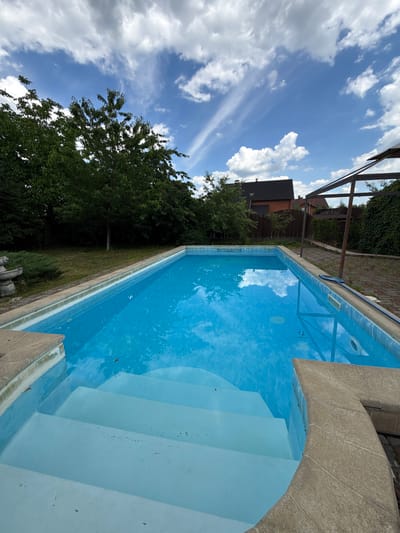 A swimming pool is pictured from a high angle, with the camera tilted. The pool is filled with clear, blue water and has a light blue staircase leading into it. The pool is surrounded by a concrete edge and a paved area. In the background, there are trees, houses, and a blue sky with white clouds. The image appears to be taken on a sunny day, with the sky reflecting in the water. There are no people visible in the image.