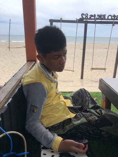 A young male with dark hair sits on a bench at a beachside cafe. He is wearing a yellow shirt with grey sleeves, and a patterned cloth draped over his legs. The beach has sand and the ocean is visible in the background, along with two swings hanging from a wooden frame. The cafe appears to be made of wood and has a roof. The overall scene suggests a relaxed atmosphere by the sea in Keudekruenggeukueh, Indonesia.