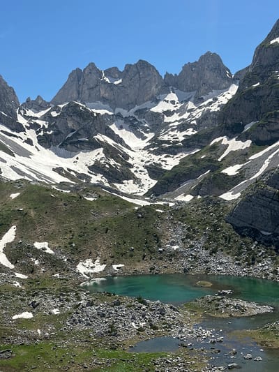 The image depicts a high-altitude mountainous landscape in Shalë, Albania, under a clear blue sky. Prominent in the upper two-thirds are rugged, grey rocky mountain peaks, extensively covered with patches of white snow, indicating a snow line. The snow is distributed irregularly, clinging to crevices and lower slopes, with exposed rock faces showing varied geological textures. Below the major peaks, the terrain transitions to steep, vegetated slopes, which are a mix of green grass and scattered rocks, interspersed with smaller snow patches and scree fields.

In the lower third of the image, a vivid turquoise-colored alpine lake is situated amidst the rocky and green terrain. The lake has an irregular shape and appears relatively shallow in areas, revealing a rocky bottom and small, rocky islets within its clear water. Adjacent to and below the main lake are smaller, interconnected pools or marshy areas with a less intense greenish hue. The immediate foreground consists of rocky ground with sparse green vegetation and a few small, residual snow patches. No individuals, animals, structures, or artificial objects are discernible within the frame. The lighting suggests a bright, sunny day, with clear shadows cast by the mountains and terrain features.