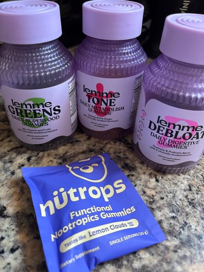 The image shows three bottles of "Lemme" brand gummy supplements, and a blue packet of "Nütrops" functional nootropics gummies, arranged on a speckled countertop. The Lemme bottles, each with a light purple lid, are partially visible. The top bottle appears to be "Lemme Debloat Daily Digestive Gummies." The middle bottle is "Lemme Tone Daily Metabolism Gummies." The bottom bottle is "Lemme Greens Daily Superfood Gummies." The Nütrops packet is labeled as "Functional Nootropics Gummies," lemon clouds flavor. The location is not discernible from the image.
