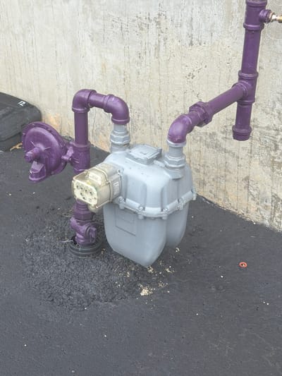 An exterior, eye-level, medium shot captures a gas meter and associated piping against a concrete wall and dark asphalt ground. The gas meter, a grey rectangular unit with a clear dial cover, is situated centrally. Purple painted pipes connect to the meter from the left and right, with a vertical purple pipe extending upwards from the right side. A circular purple component, possibly a regulator, is attached to the piping on the left. The concrete wall behind the meter exhibits a weathered, textured surface with patches of discoloration. The ground is covered in dark asphalt with scattered small white specks and some lighter-colored debris. A black bin with the number "12" visible on its side is partially visible on the far left of the frame. The scene is located in Murfreesboro, United States of America.
