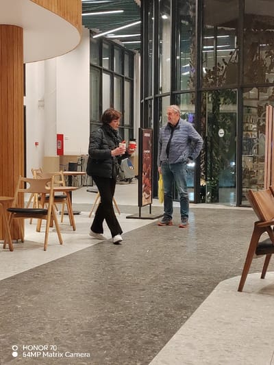 An interior view shows a woman in a black coat and pants holding two red and white cups, walking towards the camera. A man in a blue jacket and jeans stands nearby, facing her. They are in a public space with tables and chairs, next to large windows. A promotional sign stands between them. The floor is a speckled gray. A wooden pillar and various architectural elements frame the background. A watermark is visible at the bottom of the image.