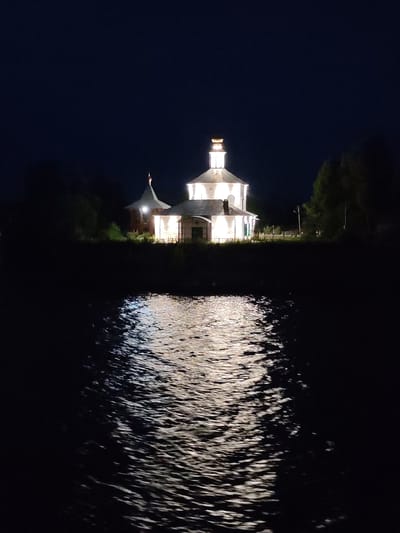 A night-time, low-angle shot captures a illuminated church with multiple domes and a central spire reflecting on a dark, rippling body of water. The church is white with a gray roof and appears to be made of wood, with a distinct, small golden dome atop the central spire and another on a smaller, adjacent dome. The water in the foreground is dark with dynamic reflections of the church's lights, creating a textured, shimmering effect. Trees and foliage are visible in the background, silhouetted against the dark night sky. The image appears to be taken from a boat or the water's edge. This scene is the Church of St. Nicholas the Wonderworker on the Yamskaya Sloboda, located in Uglich, Russia.
