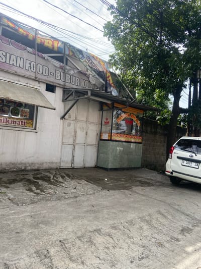 A street-level view of a small, white building in Baleendah, Indonesia, with a partially collapsed awning and a sign reading "Asian Food- Bubur Kompl". Next to the building is a smaller food stall with a sign that reads "Pondok Minang Masakan Padang". A white car with a license plate reading "D 1697 ZA" is parked in front of the stall. The scene is on a paved street in front of the building. The buildings are surrounded by overgrown vegetation.  The image is taken from the street level looking toward the buildings. The sun is shining, and the sky is clear. The street is empty, with no people in sight.