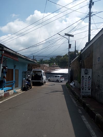 The image shows a narrow street in an urban area. On the left, a light blue building with closed doors and a blue bench sits next to a parked blue motorcycle and a white van with its back door open. In the background, a white car is partially visible. Above, there is a complex network of overhead wires. On the right, a high wall runs along the street, with a barbershop sign displayed on it. The street surface is asphalt. The sky is light blue with white clouds. A utility pole stands next to the wall. There is no evidence to pinpoint the specific location as Zeyathiri District, Myanmar.