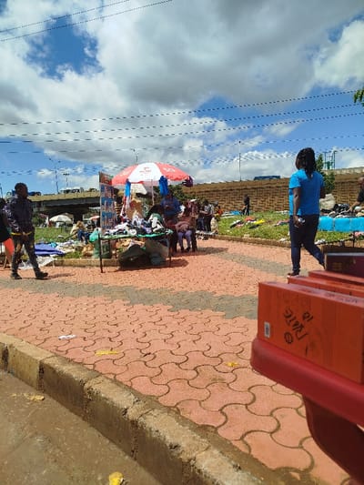The image shows a street market in Gitaru, Kenya, located at coordinates -1.232 latitude and 36.676 longitude. The market is set up on a paved sidewalk with red and grey interlocking pavers. A red and white striped umbrella provides shade for a stall selling assorted goods. Several people are present. There are four people sitting at a table underneath the umbrella. A woman in a blue shirt is walking in the center of the image, away from the camera. Two men are walking on the sidewalk, one is facing the camera and one is walking away. Another person is partially obscured by the camera, walking on the sidewalk toward the left side of the image. Several other people are visible in the background, including a person sitting on the edge of the sidewalk. There are several boxes, likely containing the merchandise for sale, stacked in the foreground. The image is taken from a low angle, creating a perspective that emphasizes the distance between the camera and the market. The sky is blue with white clouds. There are several power lines across the top of the image. A concrete wall is visible in the background, and a road and bridge are in the distance. A green grassy area is visible on the other side of the sidewalk. There are several small trees behind the wall.