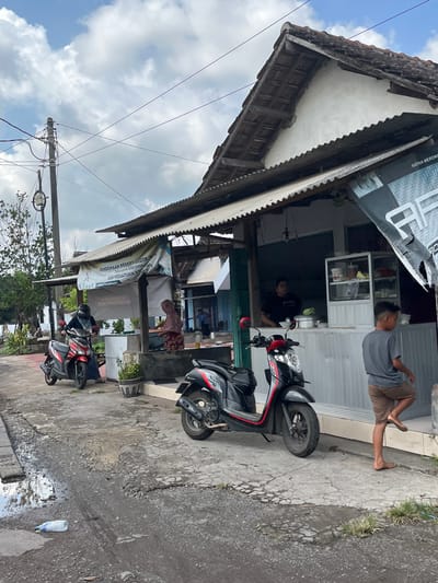 This media file depicts an outdoor scene in Jati Alun Alun, East Java. The primary subjects are two individuals, seemingly a man and a woman, engaged in activities related to a small roadside establishment. The setting appears to be a storefront or a stall with a corrugated metal roof and wooden supports.

The man is seated on the ground, leaning against a wall, wearing a grey t-shirt and brown shorts. His posture suggests relaxation. The woman is inside the establishment, tending to something on a counter. She wears a red and white patterned shirt. Two motorcycles are parked in front of the establishment, one black and red, the other red and black, indicating transportation is a common element of the scene.

The time of day appears to be daytime, with bright natural light illuminating the scene, casting subtle shadows. The sky is visible with scattered clouds, suggesting fair weather. The ground is paved and shows signs of wear and moisture, with a puddle near a plastic bottle on the right side of the image.

Notable details include the presence of plants in pots near the establishment, adding a touch of greenery. Several banners and signs are displayed, though much of the text is not clearly legible due to the image quality and orientation. One banner in the foreground has some text in Indonesian, including "PERMAINAN DESAI" and "SKP PERS". Another sign above the establishment shows "KIMIA" and other unreadable text. The overall atmosphere seems to be one of everyday life and local commerce.