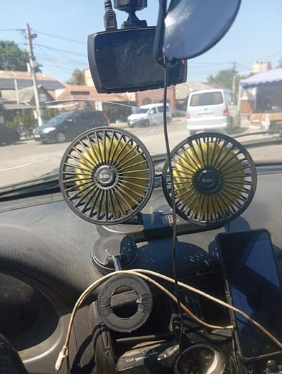This media file depicts the interior of a car, looking out through the windshield on a sunny day. The dashboard is visible, featuring two small, yellow electric fans with black grilles, mounted to the windshield. A dashcam is mounted above the fans. A smartphone in a black case rests on the dashboard to the right. Wires and a suction cup mount are also visible on the dashboard. Outside the car, a street scene is visible with buildings, trees, power lines, and other vehicles, including a white van and a black car, suggesting it is daytime. The sky is clear and blue. The scene is likely in Vasylkiv, Ukraine. No people are visible in the image.