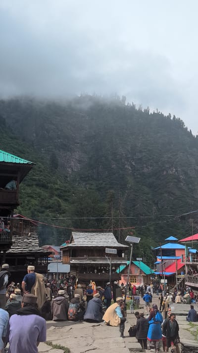 The image depicts a lively gathering in Malana, India, with a large crowd of people participating in what appears to be a festival or communal event. The scene is set against a backdrop of traditional wooden architecture and a lush green mountainside shrouded in mist. Many individuals are wearing traditional attire, including woolen shawls and caps, and are seated or standing in the central plaza. Some are engaged in conversation, while others observe the activities around them. In the background, colorful buildings and a prominent solar panel suggest a blend of traditional and modern elements. The overall atmosphere is one of community and cultural celebration, with the imposing mountain range adding a sense of natural grandeur to the setting.
