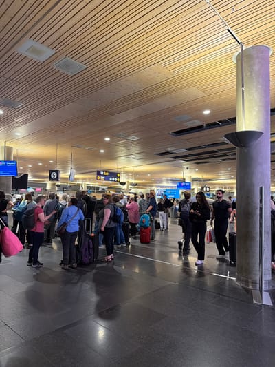 This is a photograph taken at Oslo Airport Gardermoen, Norway, depicting a queue of people with luggage in an indoor terminal. The ceiling is constructed of wooden slats, and there are various signs and lighting fixtures visible. In the foreground, people are standing in line, some with suitcases and backpacks. The floor is a dark, polished stone. In the mid-ground, there are directional signs, including one indicating "Ut Exit Toilets." To the right, a large concrete pillar with a metallic base is prominent. The overall atmosphere suggests a busy travel hub.