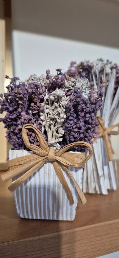 The image shows two bouquets of dried lavender in decorative pots. The pot in the foreground is white with vertical ribbing and tied with a raffia bow. The second pot, slightly behind and to the right, also appears to be white and ribbed, with a raffia tie visible. The lavender in the foreground bouquet is a deep purple, interspersed with some lighter, dried stems. The lavender in the background bouquet is less distinct due to the depth of field but appears to be similar in color and type. The objects are placed on a wooden surface, possibly a shelf or table. The background is out of focus, showing a gradient of light and shadow against a textured surface, likely a wall. There are no people or animals present in the image. The scene is static and appears to be an indoor arrangement.